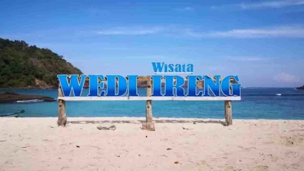 A beach with a sign reading 'Wisata WEDI TRENG' against a backdrop of a clear blue sky and calm sea. 1