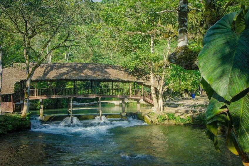 A wooden bridge over a flowing river, surrounded by lush greenery and a thatched-roof structure. 1