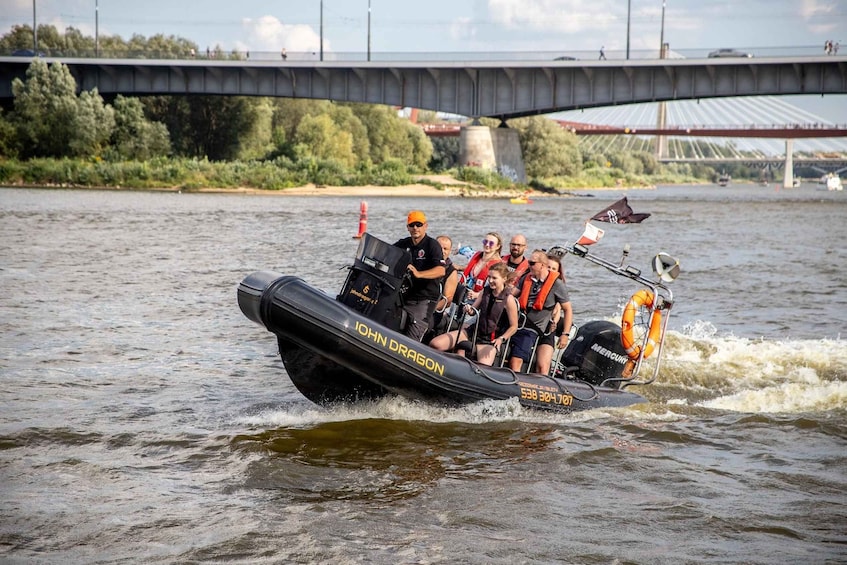 A group of people on a speedboat with 'JOHN DRAGON' written on it, moving across a river with a bridge in the background. 3