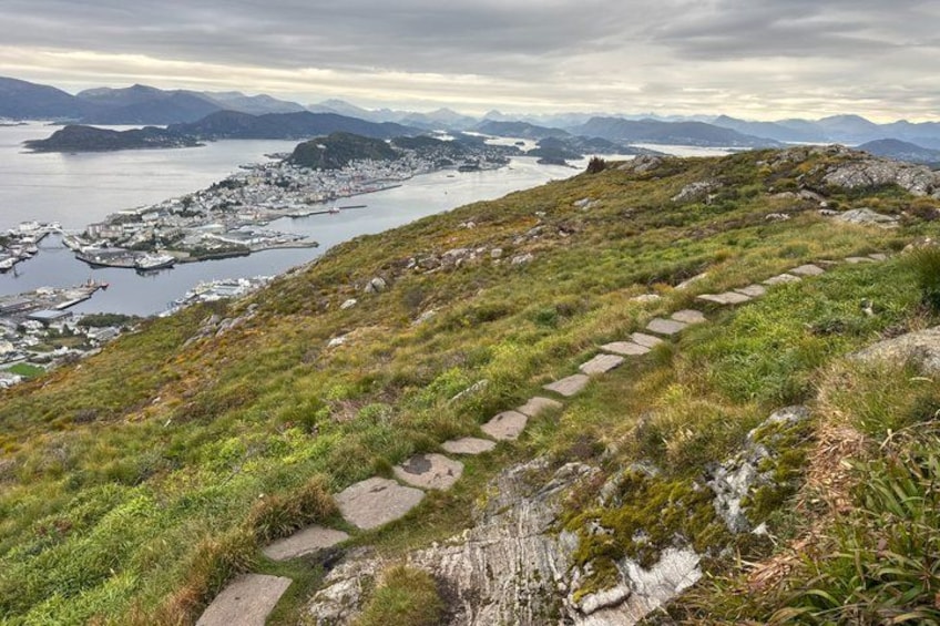 A coastal landscape with a stone pathway, grassy terrain, and a distant view of a town and water. 5