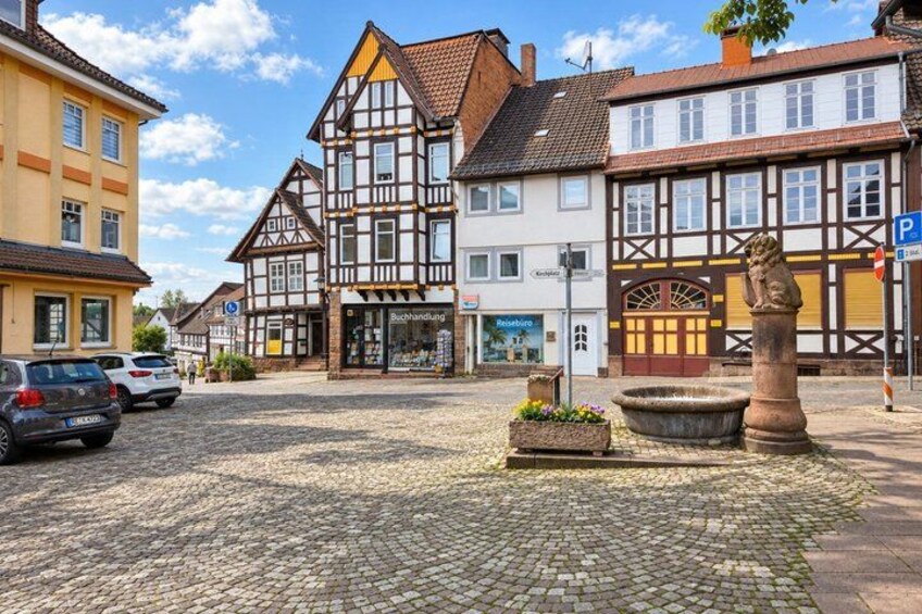A cobblestone street with traditional half-timbered buildings, a stone fountain, and a parked car. 3