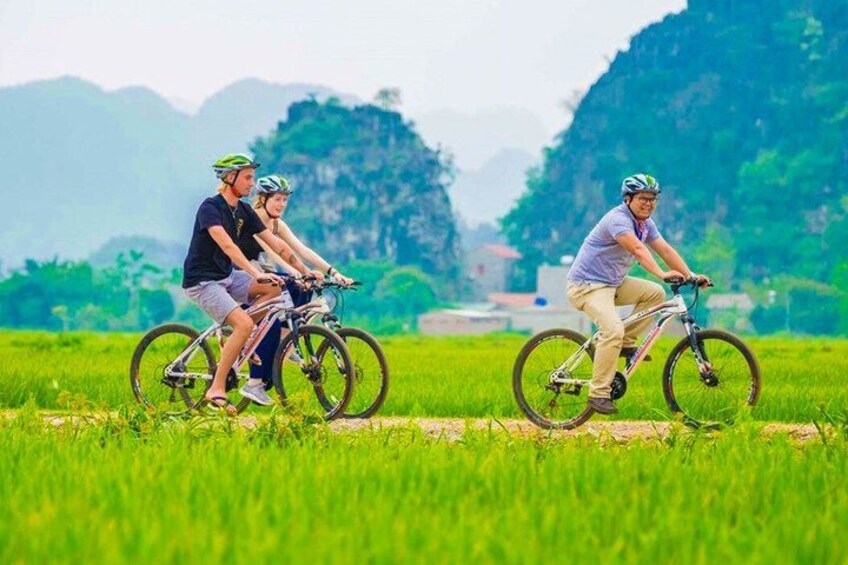 Three people riding bicycles on a path through a green field with mountains in the background. 4