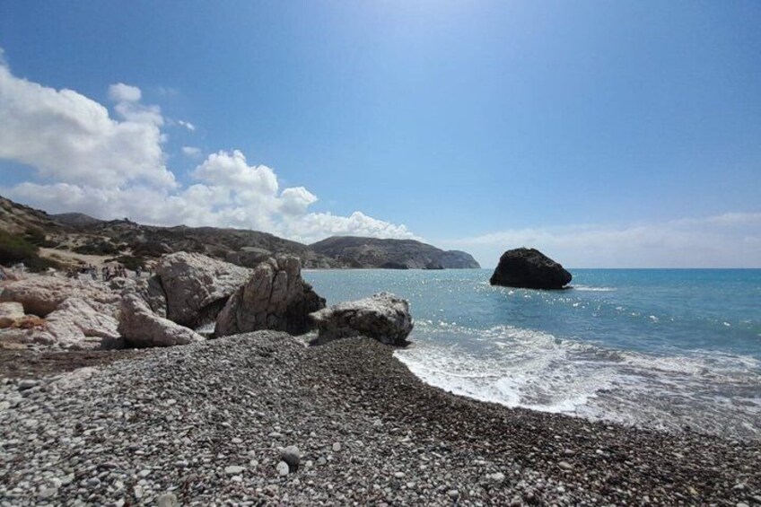 A pebble beach with large rocks, calm sea, and distant hills. 1