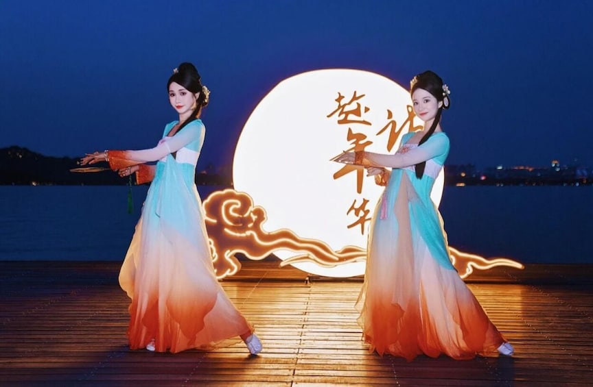 Two individuals in flowing dresses performing a dance on a wooden deck with a large illuminated moon and Chinese characters in the background. 1