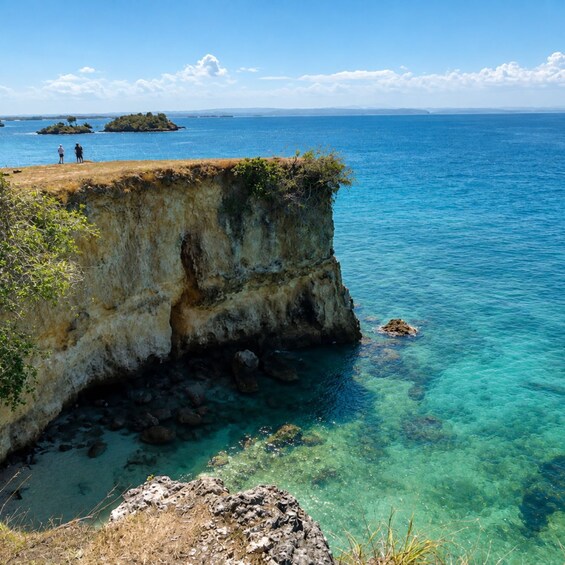 A coastal cliff with clear blue water, a rocky shoreline, and two people standing at the edge. 2