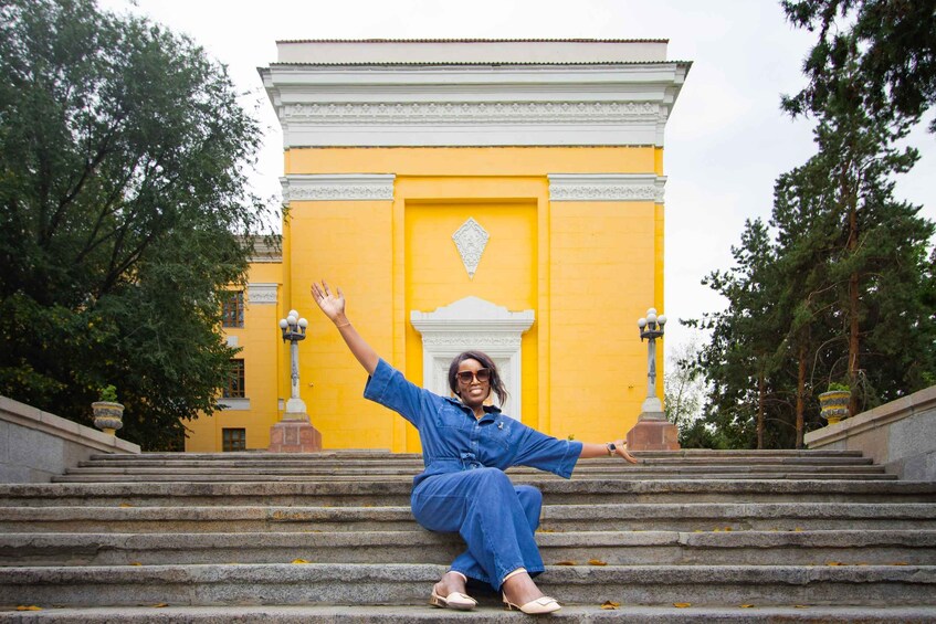 A person sitting on stone steps in front of a yellow building with white trim and decorative elements. 5