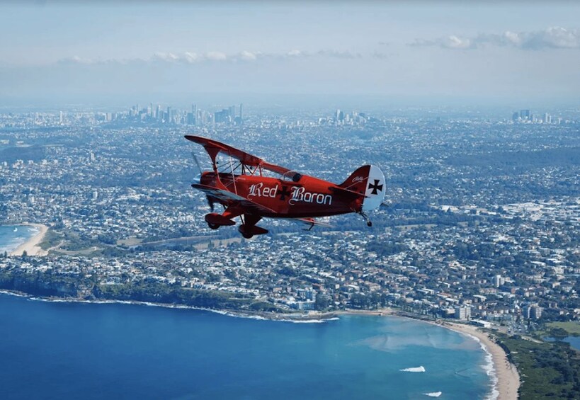 A red biplane with 'Red Baron' written on it flying over a coastal city with a clear view of the ocean and a distant city skyline. 4