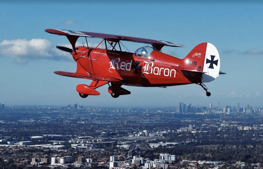 A red biplane with 'Red Baron' written on it flying over a cityscape. 1