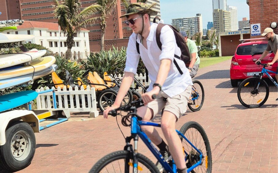 A person riding a blue bicycle on a paved path, with surfboards stacked in the background and a red car parked nearby. 4