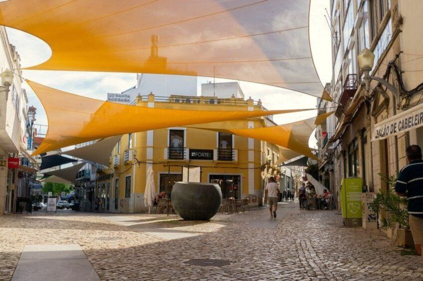 A cobblestone street with a large yellow umbrella structure, a cafe, and a building with a sign that reads 'SALÃO DE CABELIR'. 2