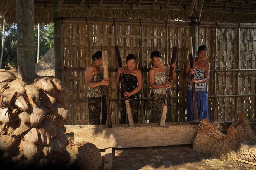 A group of people in traditional attire standing in a bamboo structure with thatched roofs, surrounded by palm fronds and coconut husks. 5
