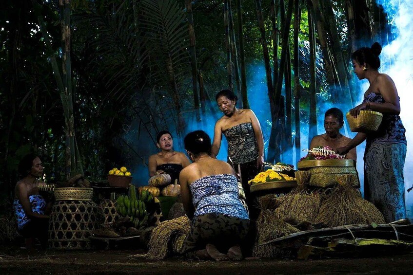 A group of people in traditional attire, gathered around a central area with baskets and fruits, set in a forested environment. 1