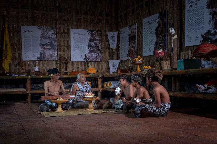 A traditional ceremony with participants seated on the floor, surrounded by wooden walls with informational posters. 3