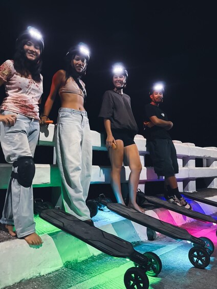 Four people standing on a platform with skateboards, under colorful lights. 2
