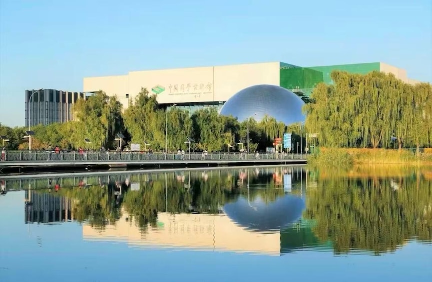 A modern building with a large dome structure, a calm body of water reflecting the scene, and a bridge with pedestrians. 1