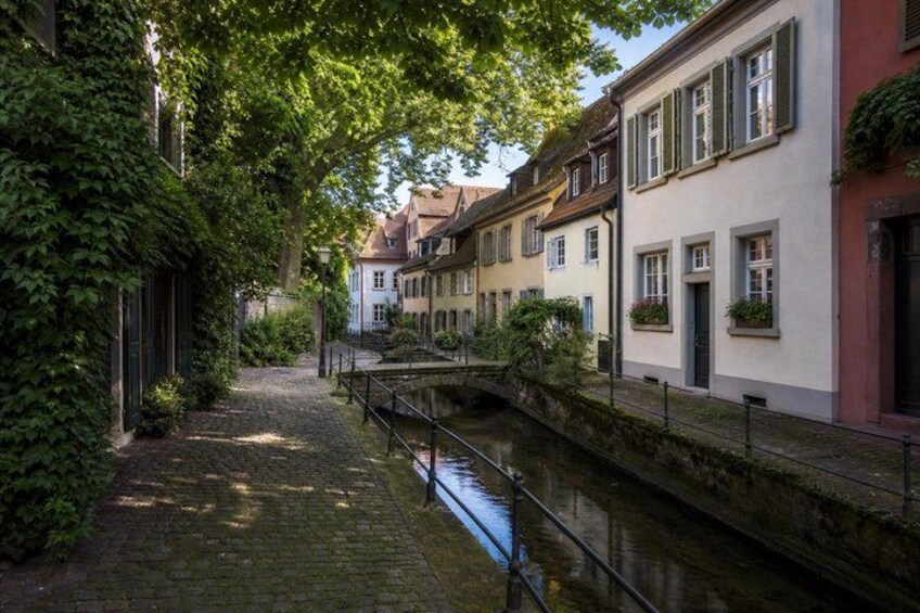 A narrow cobblestone street lined with historic buildings, a canal, and lush greenery. 3