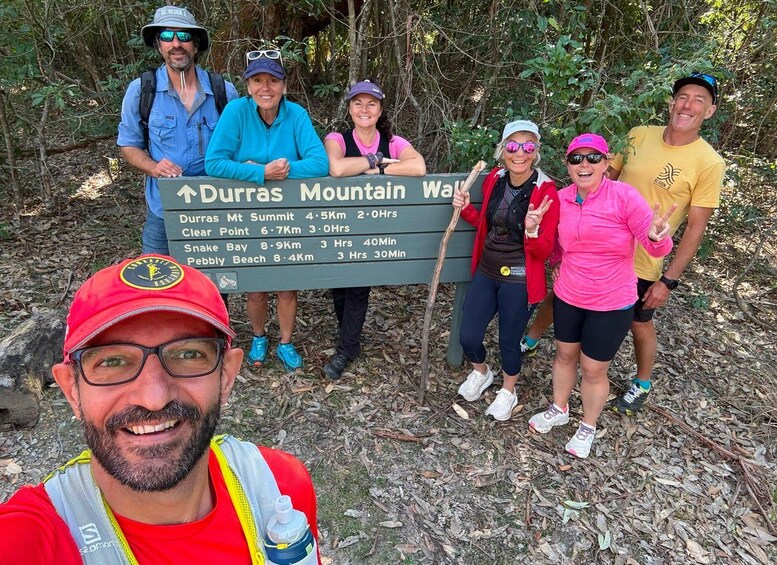 Un grupo de personas en un área boscosa, junto a un poste con indicaciones de Durras Mountain Way y varios destinos de senderismo, con distancias y tiempos. 4