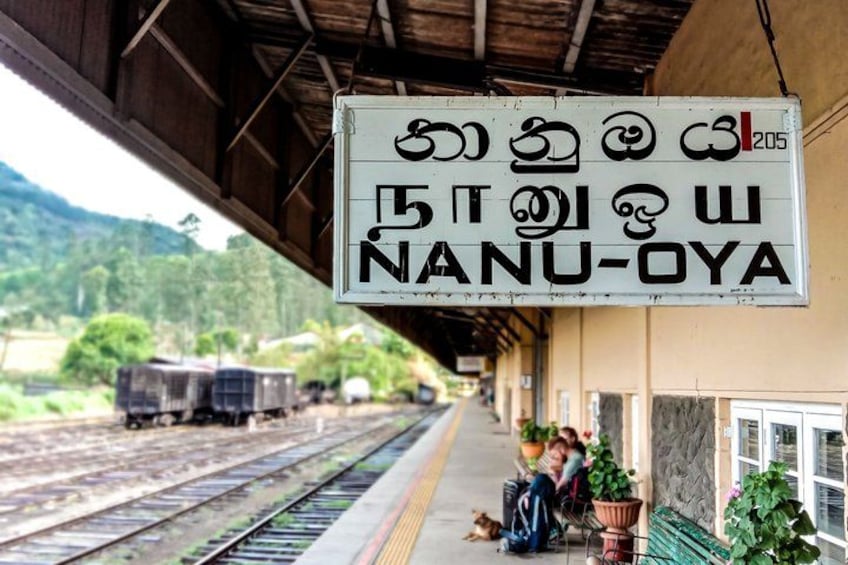 A train station platform with a sign in a local language, a train on the tracks, and a person sitting on a bench. 5