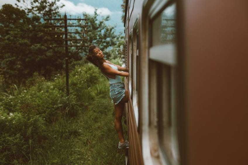 A person standing on a train, holding onto the side, with greenery and a fence in the background. 1