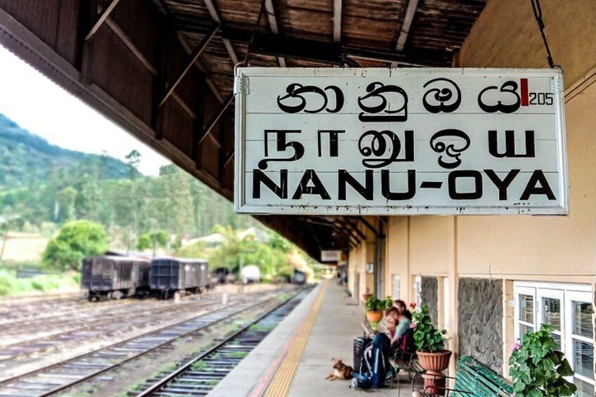 A train station platform with a sign in a local language, a train on the tracks, and a person sitting on the platform. 5