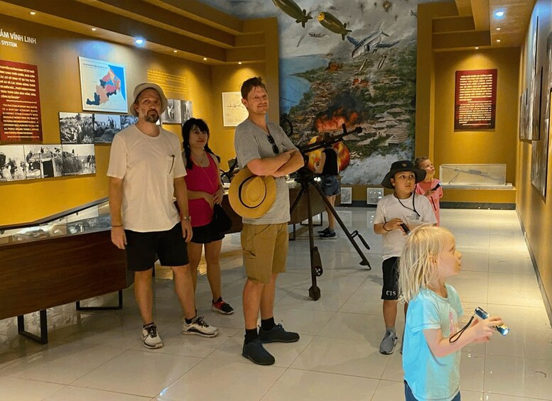 A museum exhibit with a large mural of a battle scene, informational plaques, and visitors observing the exhibit. 2