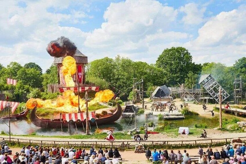 A Viking ship on fire with smoke and flames, surrounded by spectators and a small stage. 3