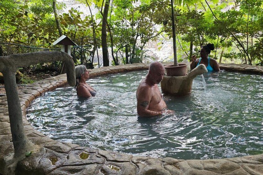 A natural hot spring with people bathing, surrounded by trees and a stone structure. 4