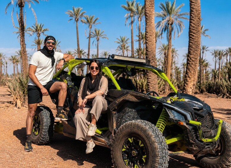 Two people sitting in a green off-road vehicle with large tires, parked in a desert with palm trees in the background. 5