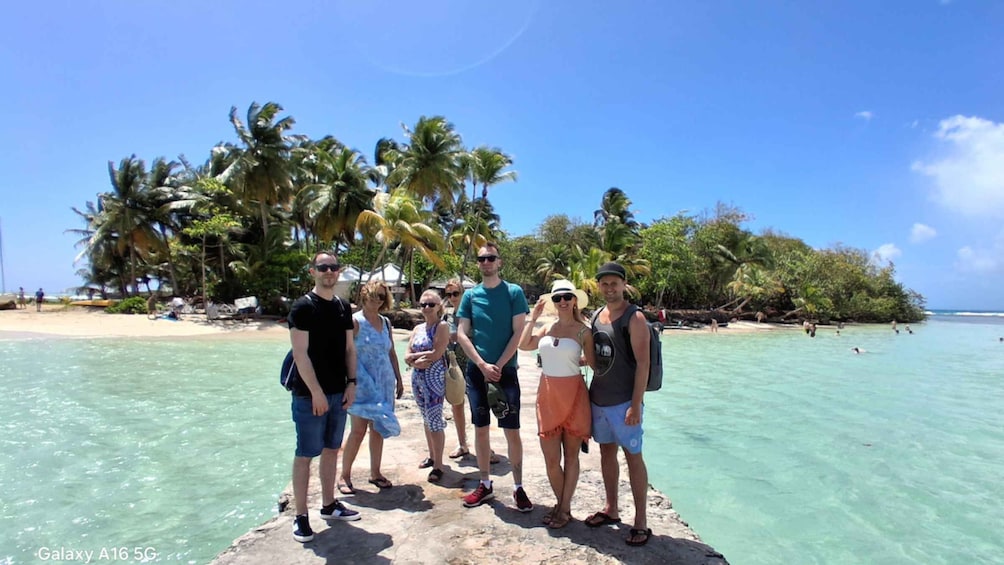 A group of people standing on a rock by a turquoise sea with palm trees in the background. 1