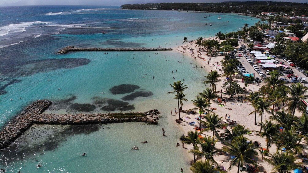 A coastal beach with clear turquoise waters, a rocky breakwater, and a densely populated area with buildings and palm trees. 2