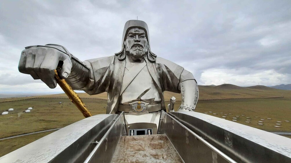 Una gran estatua de una figura con un báculo, frente a un paisaje con colinas lejanas y un cielo nublado. 4