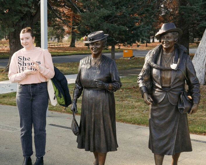 Une personne se tient à côté d’une statue représentant une femme en robe et en chapeau, avec des arbres et un banc en arrière-plan. 4