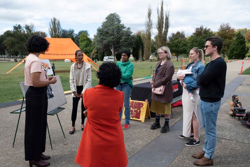 Un groupe de personnes se tient à l’extérieur, certaines tenant des pancartes. On aperçoit une tente et une pancarte sur laquelle on peut lire « CAMP OUVERT » à l’arrière-plan. 5