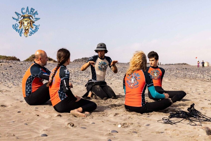 Un groupe de personnes assises sur une plage de sable, portant des combinaisons de plongée, avec un logo dans le coin supérieur gauche. 1