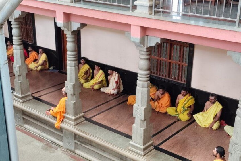 A group of monks in traditional orange robes seated on a porch with a pink building in the background. 4