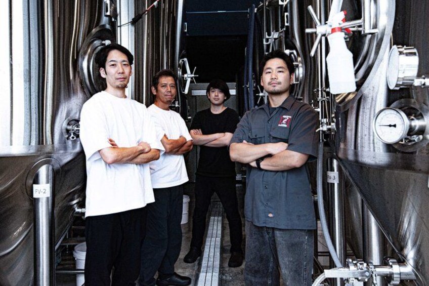 Four people standing in a brewery with stainless steel tanks and pipes. 2