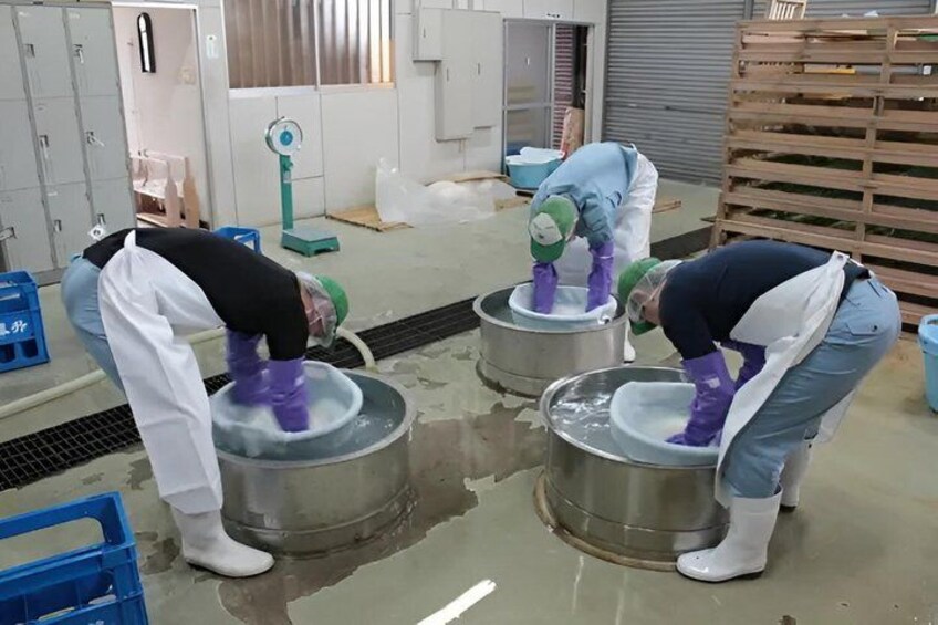 Two individuals in a workshop bending over large metal containers, likely engaged in a task involving liquids. 5