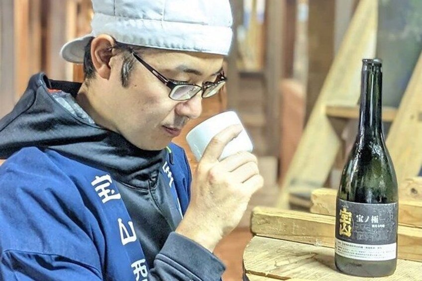 A person wearing a blue jacket with white text, sitting at a wooden table with a bottle of wine and a white cup. 2