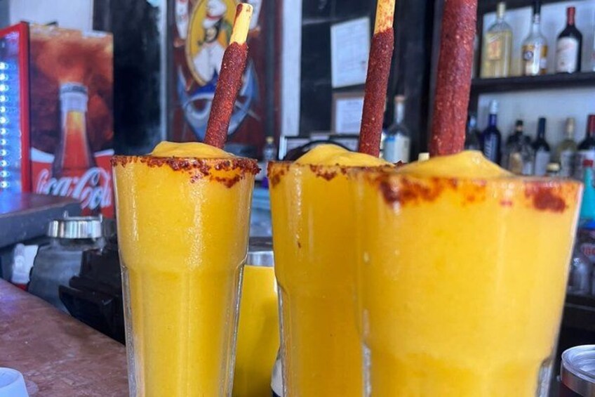 Two tall glasses of a yellow beverage with a red garnish, placed on a bar counter with a Coca-Cola can and other bottles in the background. 1