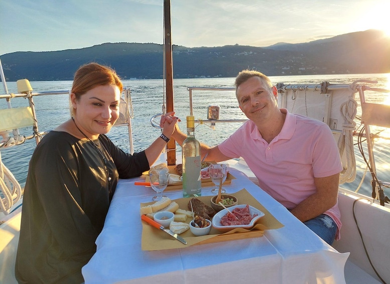 Two people sitting at a table with food and drinks on a boat with a scenic view of the water and hills in the background. 1