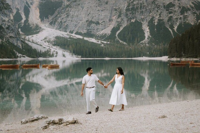 A couple holding hands on a lake shore with boats and mountains in the background. 1