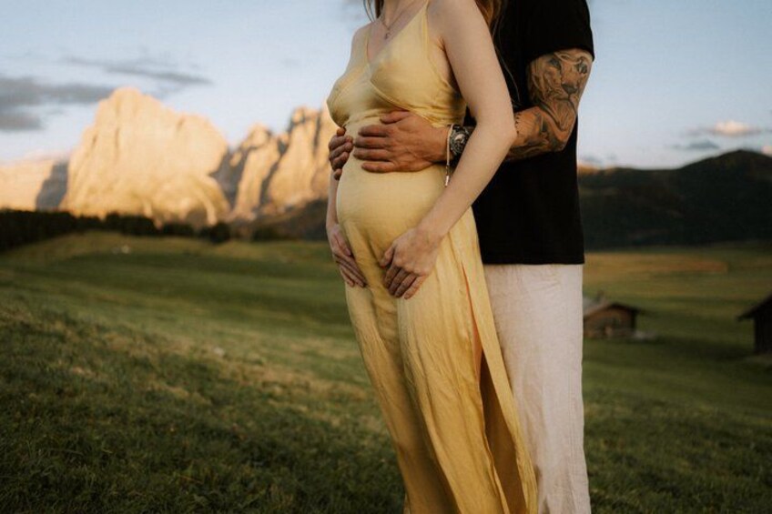 A pregnant woman in a yellow dress holding her belly, with a man standing beside her, in a grassy field with mountains in the background. 4