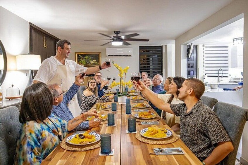 A group of people toasting at a wooden dining table with plates of food and drinks. 2