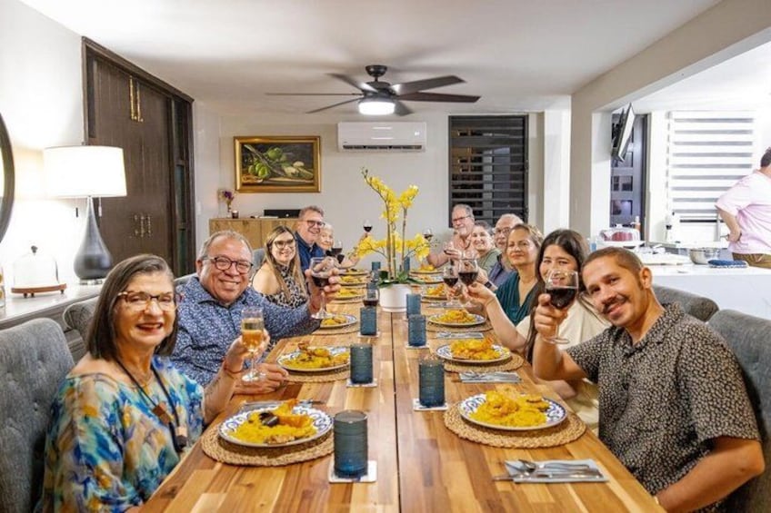 A group of people seated around a wooden dining table with plates of food and glasses of wine, in a well-lit room with a ceiling fan. 1