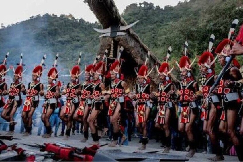 A group of people in traditional attire with red and black patterns, standing in formation with spears and shields, in a natural outdoor setting with trees and a thatched structure in the background. 2