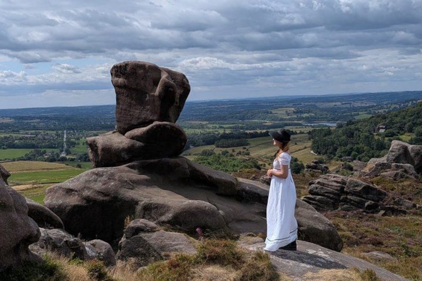 A person in a white dress standing on a rocky outcrop overlooking a valley with a large rock formation and distant landscape. 1