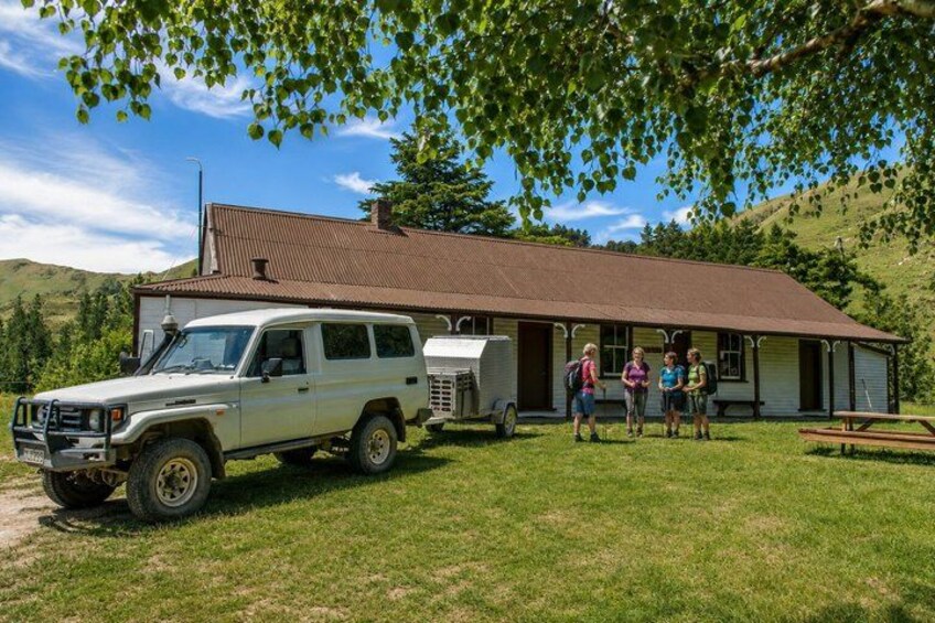 A white SUV parked in front of a rustic building with a trailer, surrounded by greenery and hills. 2
