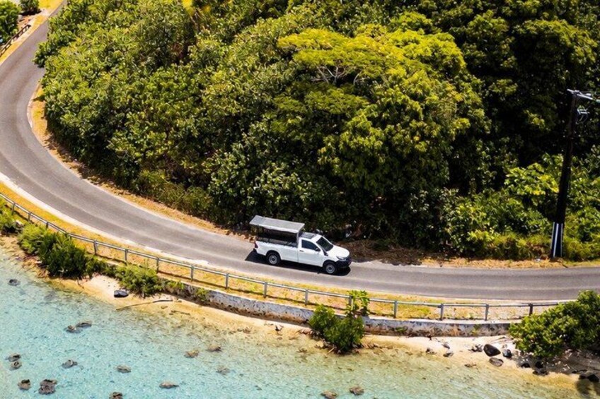 A white vehicle on a winding road surrounded by dense greenery and a beach. 1