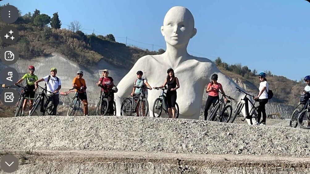A group of cyclists on a rocky path with a large white sculpture in the background. 1