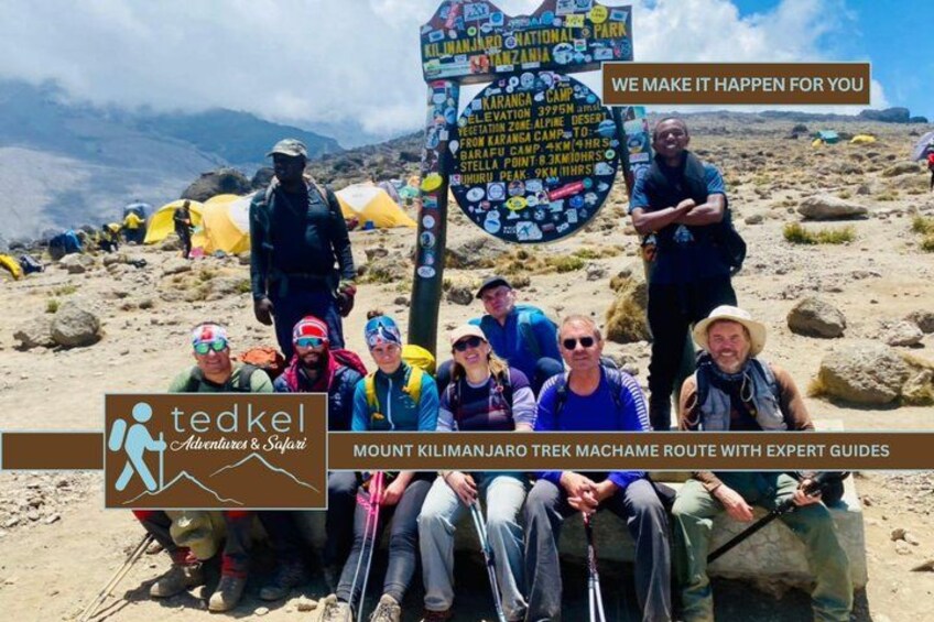A group of people at a mountain campsite with a signpost listing various camps. 1